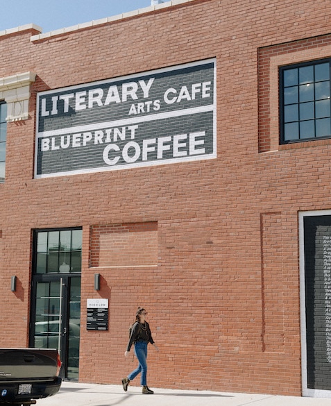Nora 5 Nora Brady walks on the sidewalk next to Blueprint Coffee High Low in front of the sign that reads "Literary Cafe"
