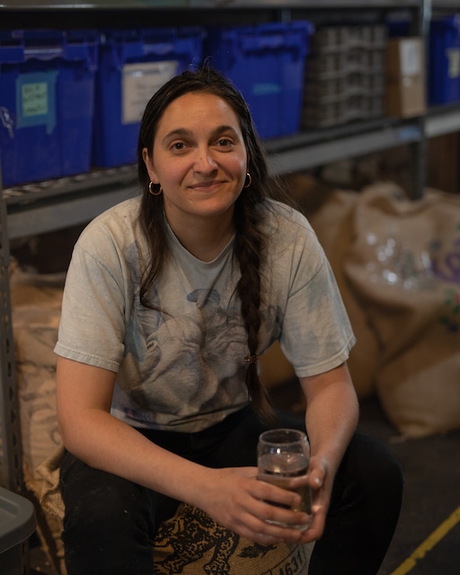 2023_05-45.jpg beaan Marija sits in front of a shelf of burlap sacks of green coffee, looking at the camera and smiling