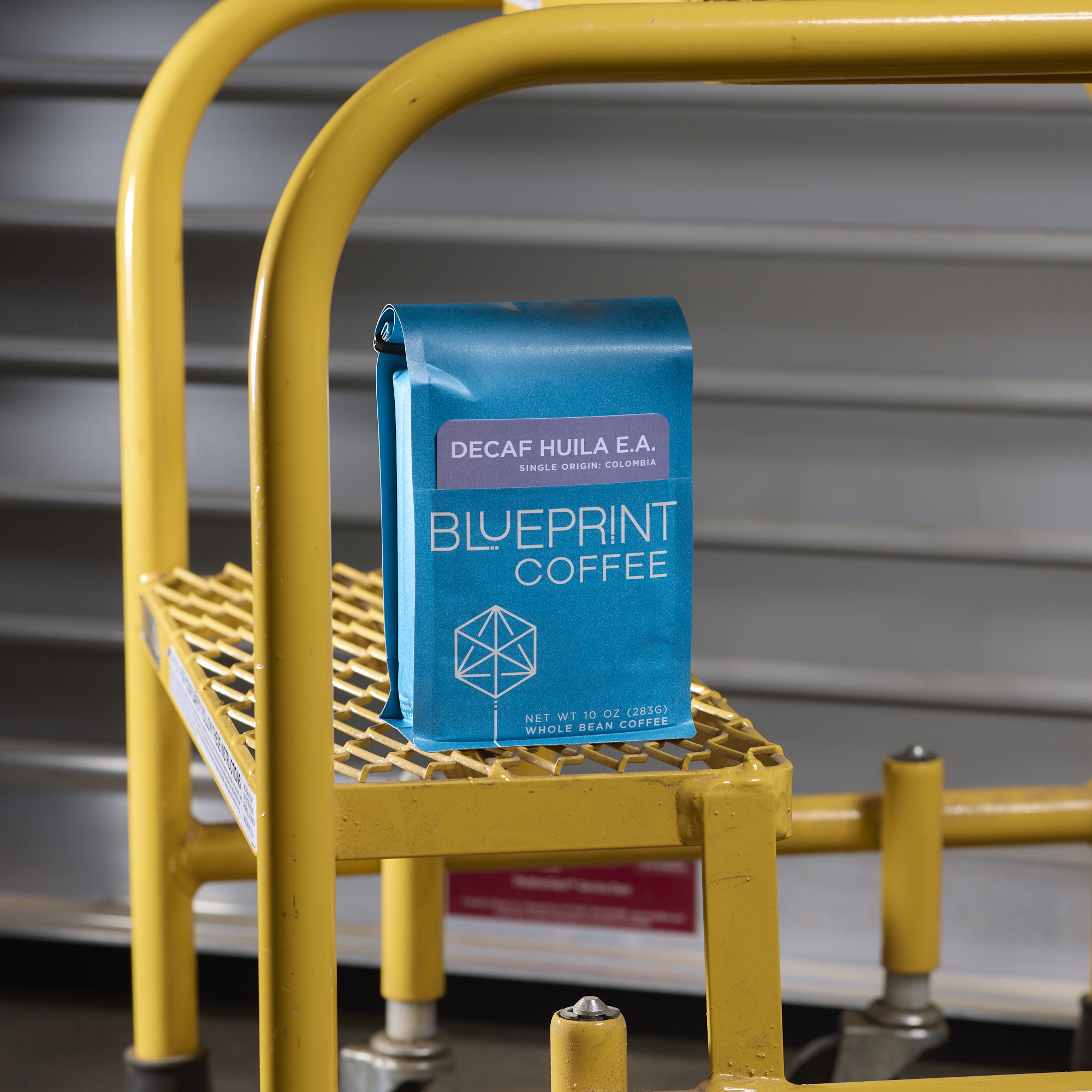 A bright blue bag of Blueprint Coffee Decaf Huila E.A. sits on the step of a yellow stepladder