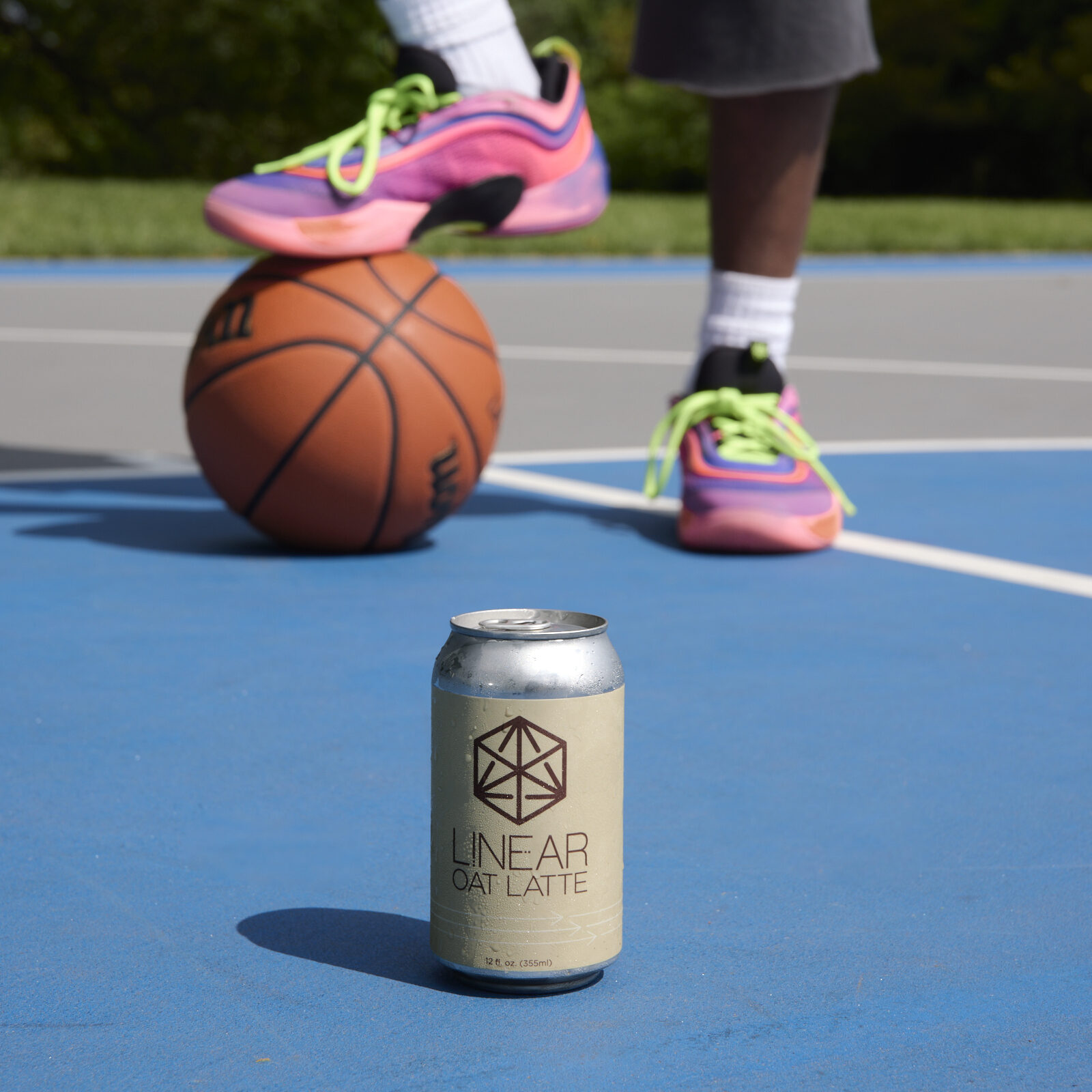 A can of Linear Oat Latte on a blue basketball court. In the background is a man wearing pink sneakers with one foot propped up on a basketball.