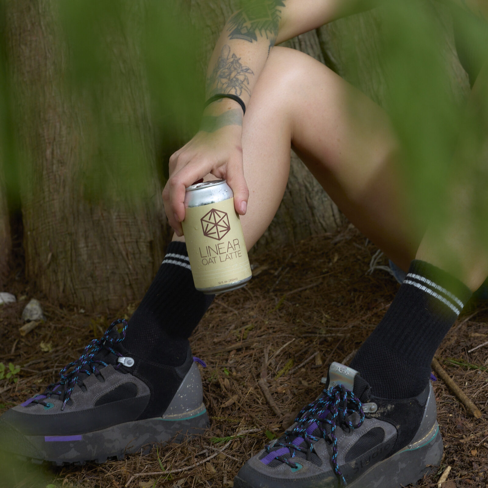 A person sitting among leafy plants outdoors hols a can of Linear Oat Latte