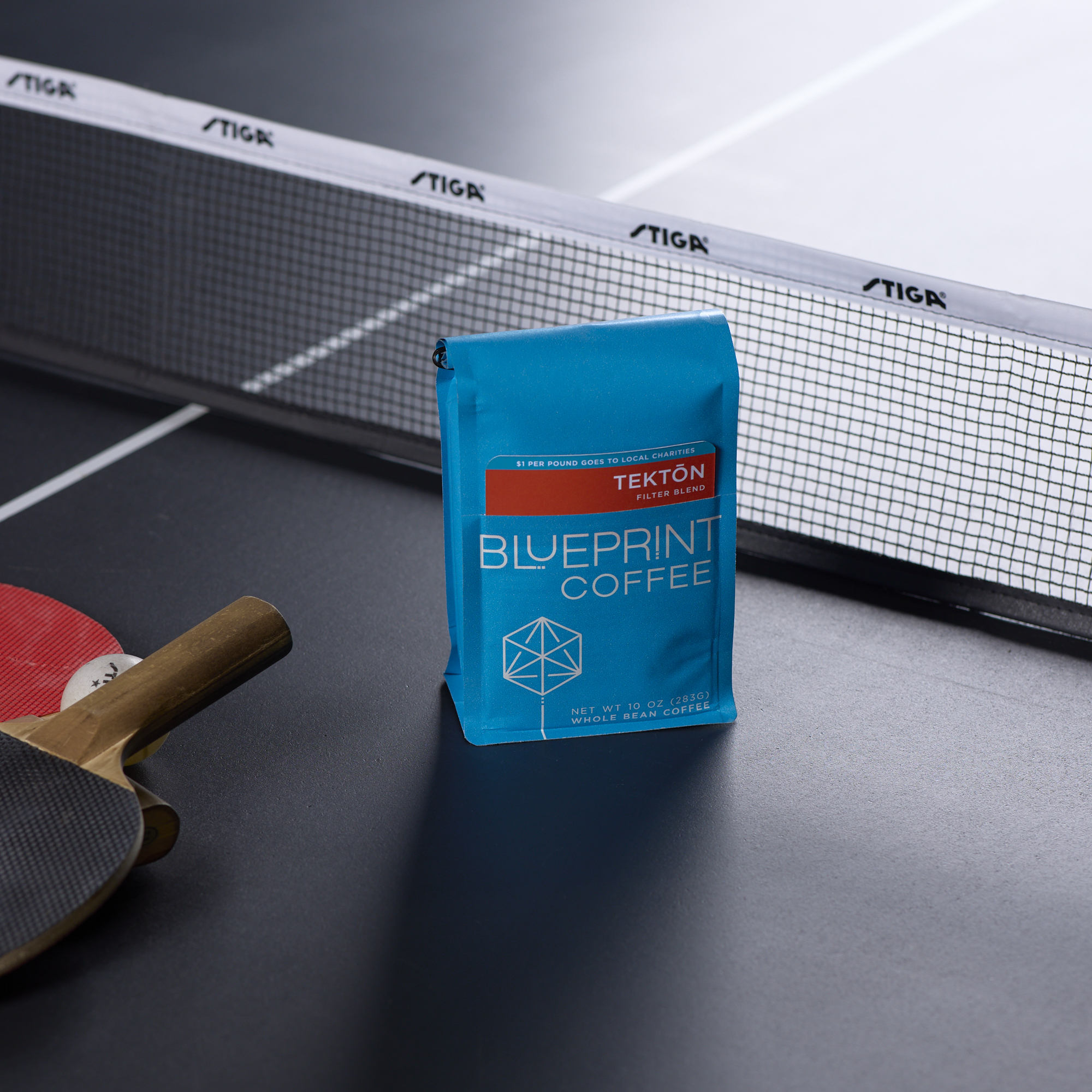 A blue 10-ounce bag of Tektōn Filter Blend stands on a table tennis table near the net, beside a paddle and ball