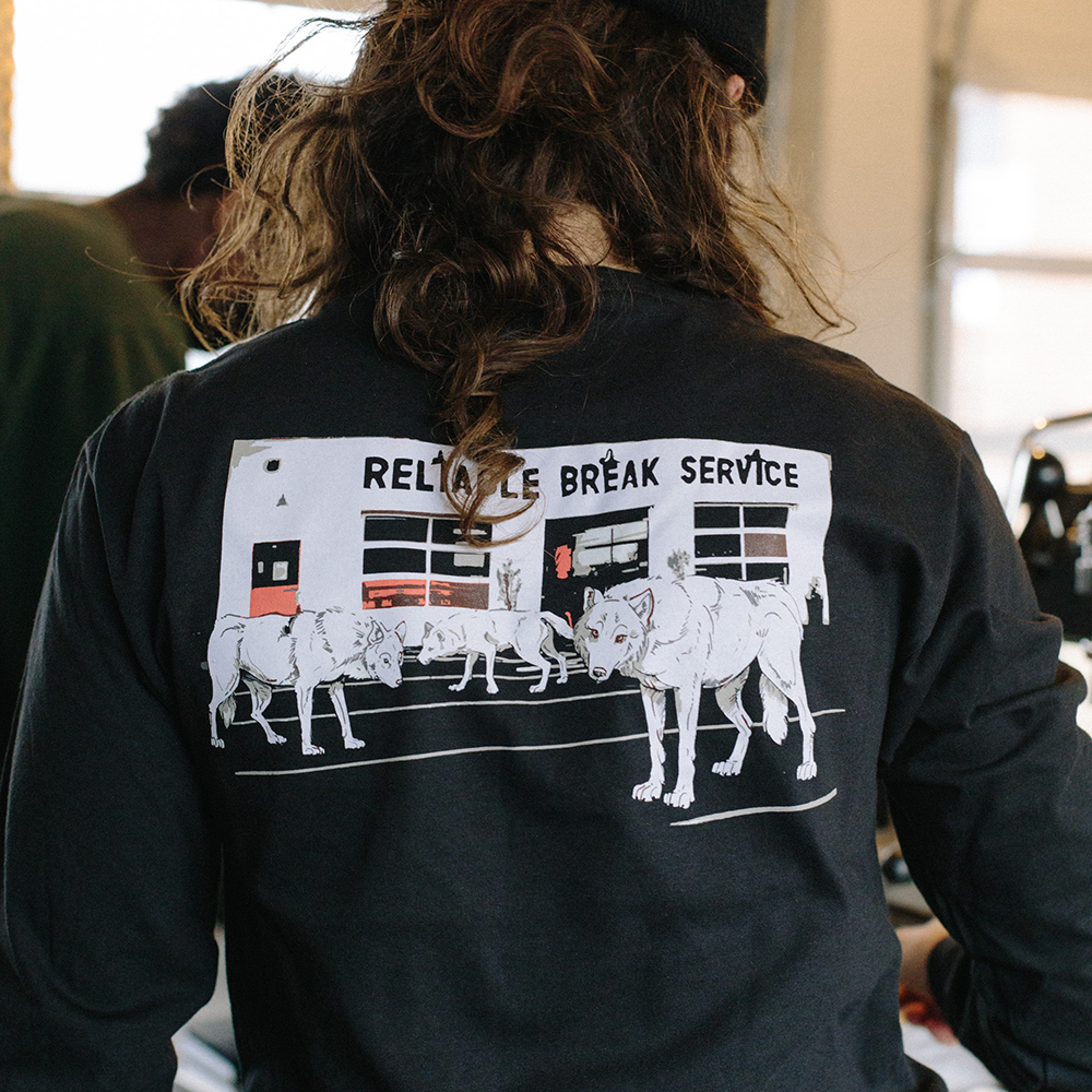 A person's back is turned to the camera, showing the screenprinted illustration printed on the back of their long-sleeve black tee: three wolves in front of our Watson coffee bar.