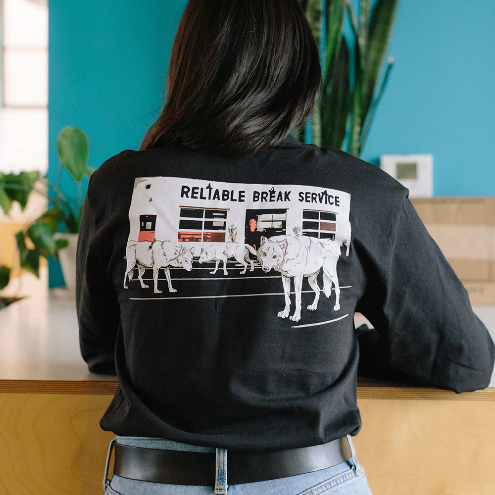 A person's back is turned to the camera, showing the screenprinted illustration printed on the back of their long-sleeve black tee: three wolves in front of our Watson coffee bar.