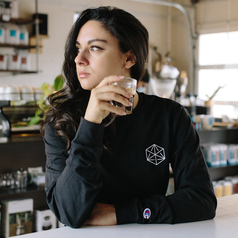 A person holds an espresso shot and looks away to the side. Their long-sleeved black t-shirt is printed on the front with a Blueprint "cube" logo.