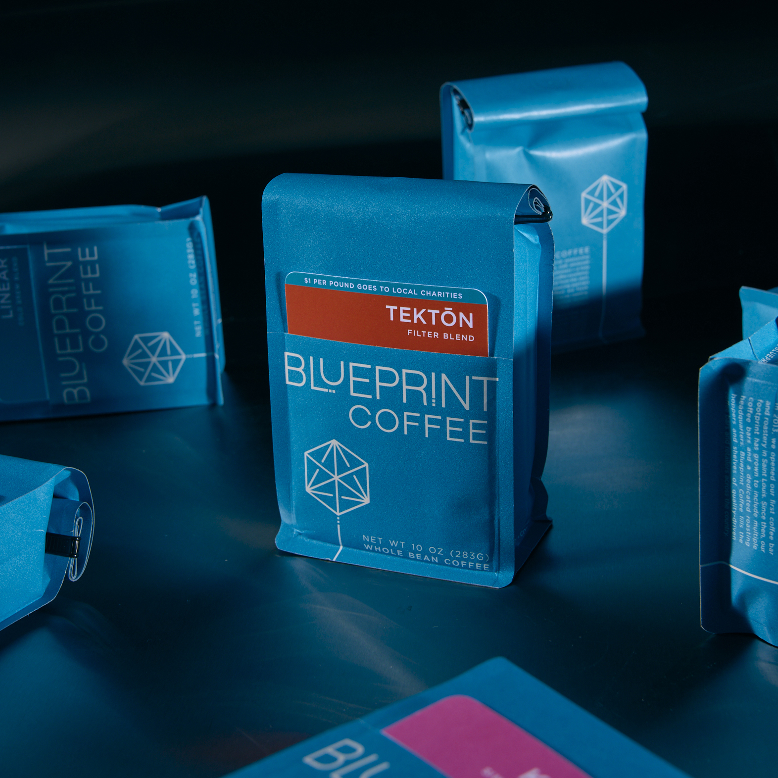 Blue bags of Blueprint Coffee beans are arranged in various positions, showing the front, back, and sides. In the center is a bag of Tektōn coffee beans, labelled with a bright orange removable card.