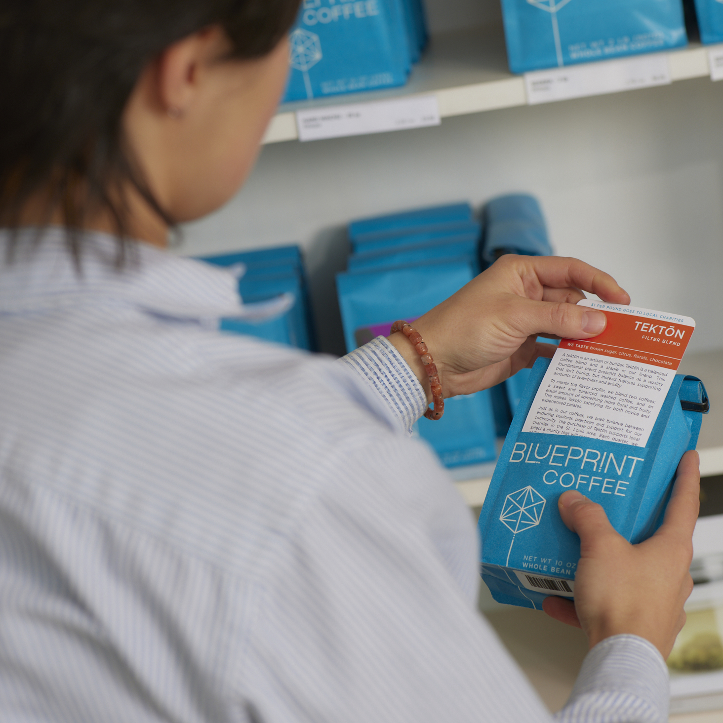 A person stands in front of a retail shelf stocked with coffees and teas. The person is holding a bag of Tektōn coffee beans; they are reading text printed on the removable label card which they've partially pulled from its pocket in the bag.