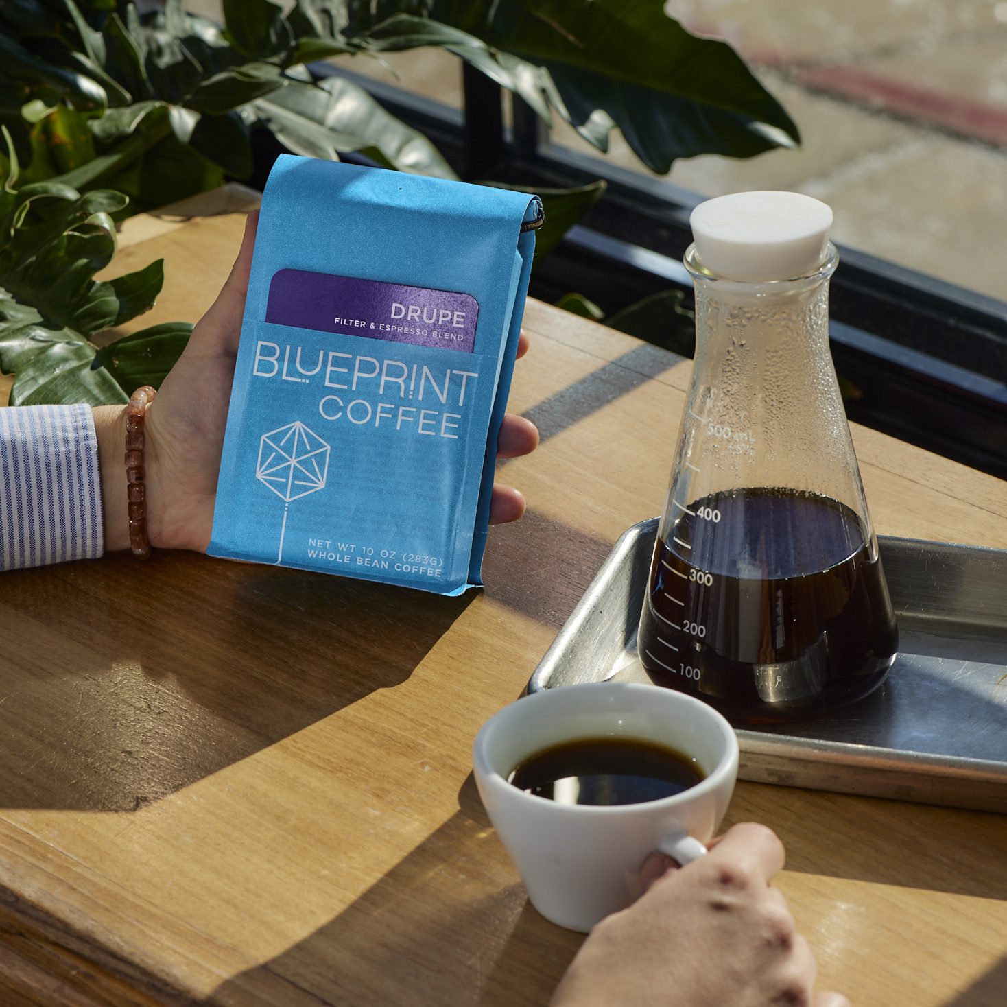 A person sits at a wooden countertop with a mug of brewed coffee in their right hand, and in their left hand is a bright blue bag of Drupe coffee beans.