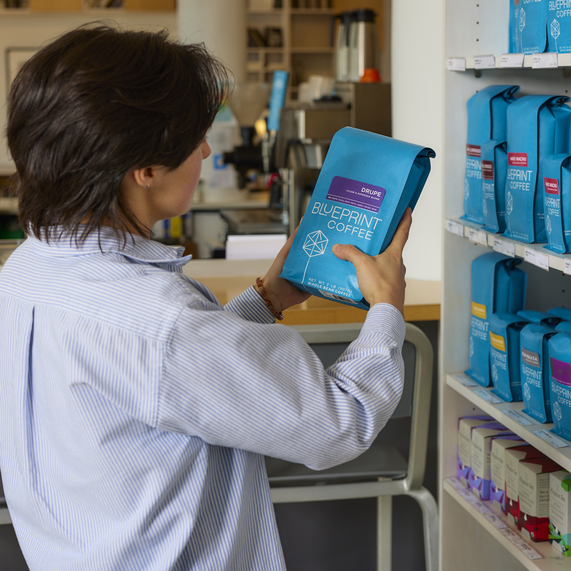 A person stands in front of a retail shelf stocked with coffees and teas. The person is holding a 2 pound bag of Drupe coffee beans.