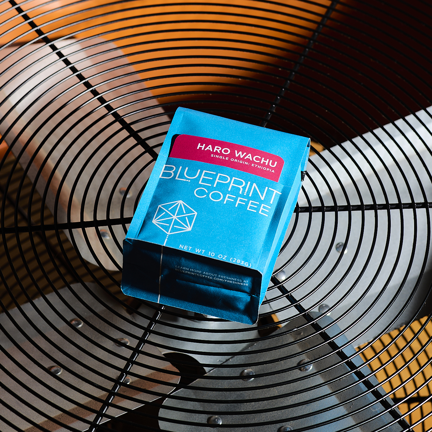 A bright blue bag of Haro Wachu, Ethiopia coffee beans with the Blueprint Coffee logo and a red removable info card. The bag is placed on a bright orange industrial fan with a radial grid pattern.