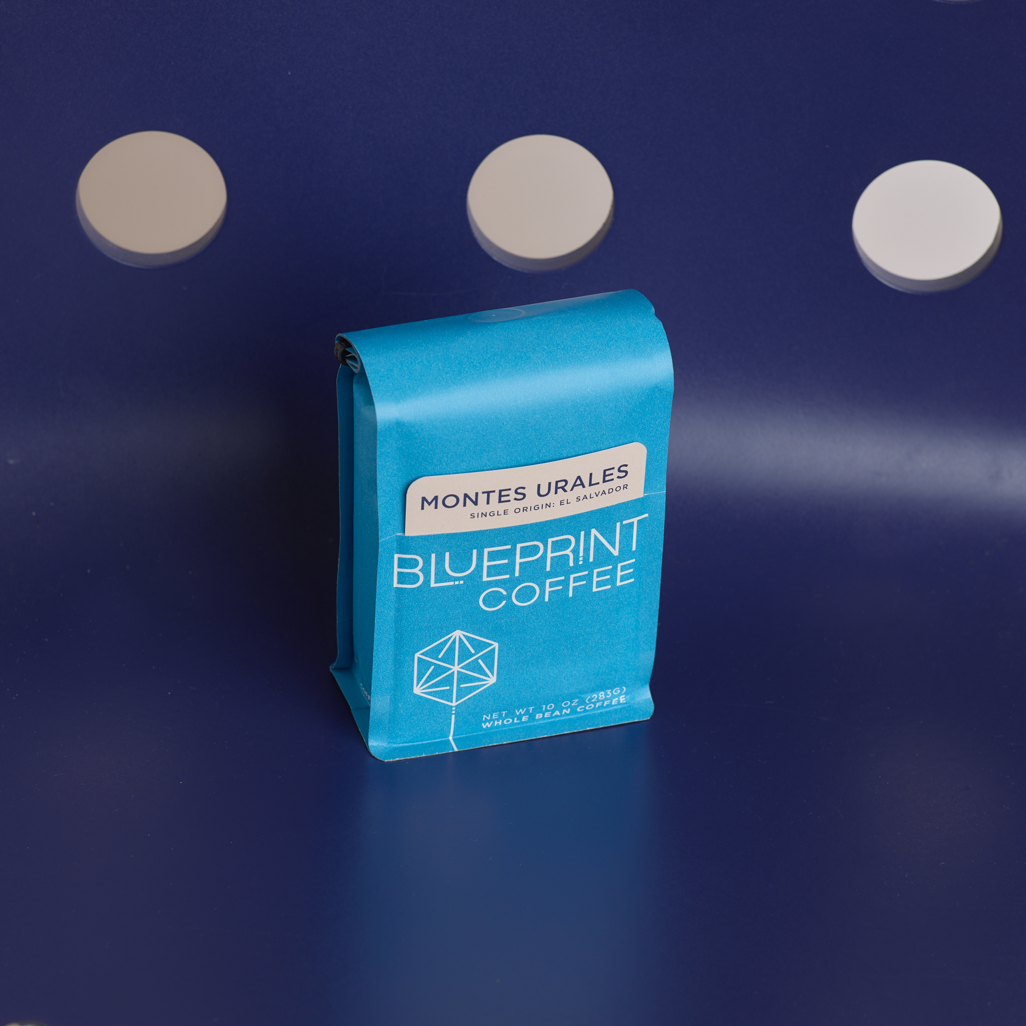 A bright blue bag of Montes Urales, El Salvador coffee beans with the Blueprint Coffee logo and a tan removable info card. The bag is centered over a dark navy surface; three round cutouts above the bag reveal a light tan background.