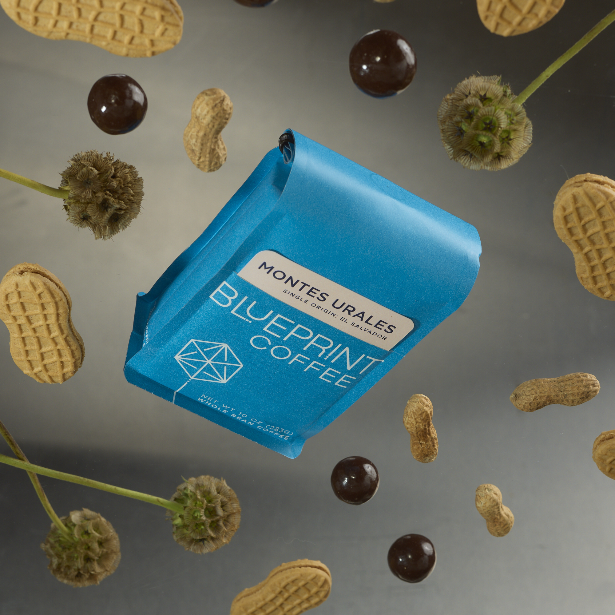 A bright blue bag of Montes Urales, El Salvador coffee beans with the Blueprint Coffee logo and a tan removable info card. The bag appears to be floating, with peanuts, Nutter Butter cookies, green flowers, and chocolate covered malt balls surrounding it.