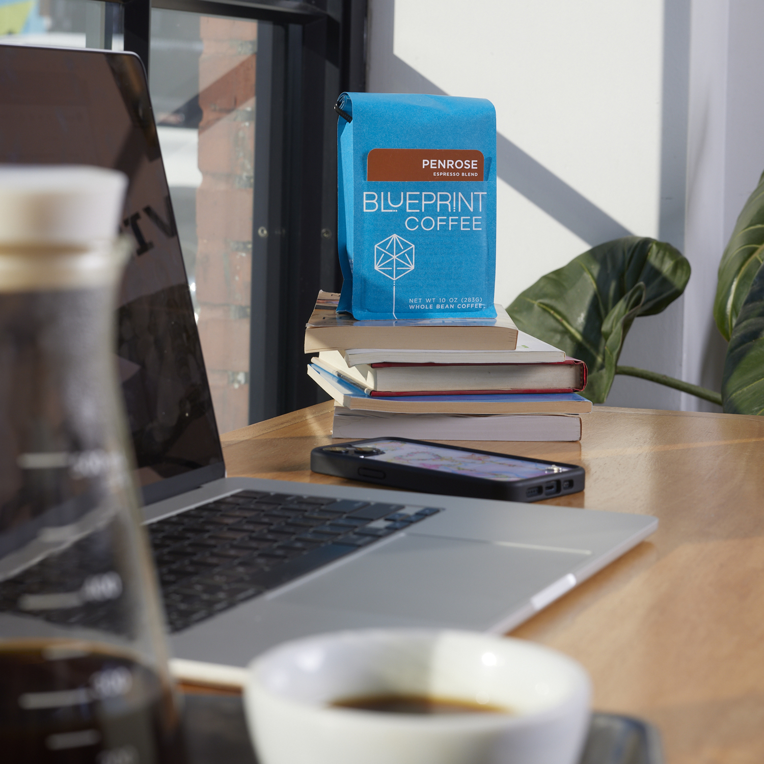 A blue bag of Penrose coffee beans is perched atop a stack of books on a wooden countertop. In the foreground is a person's open laptop and a brewed drip coffee, partially enjoyed.